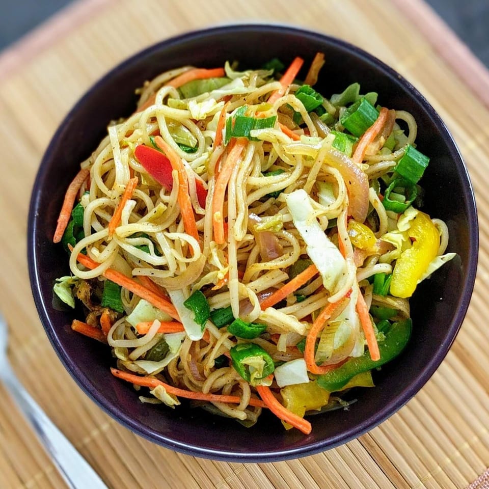 A dark bowl filled with Indian-style noodles, carrots, cabbage, onions, and green peppers on a bamboo mat.