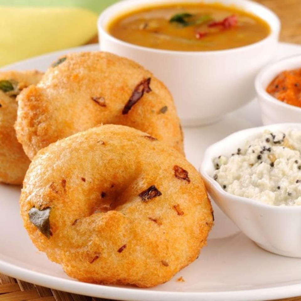 Golden-fried Indian vada donuts with sambar, coconut chutney, and red chutney on a white plate.