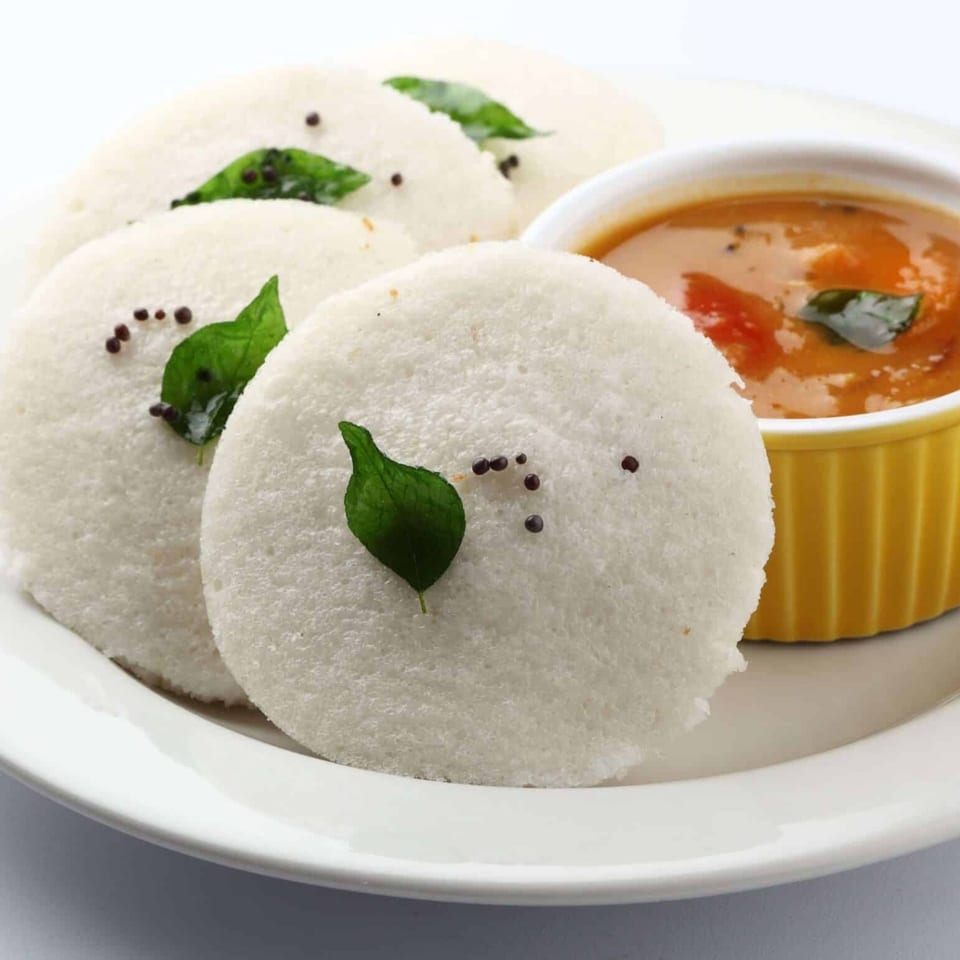 Four white idlis garnished with curry leaves and mustard seeds, served with a yellow bowl of sambar.