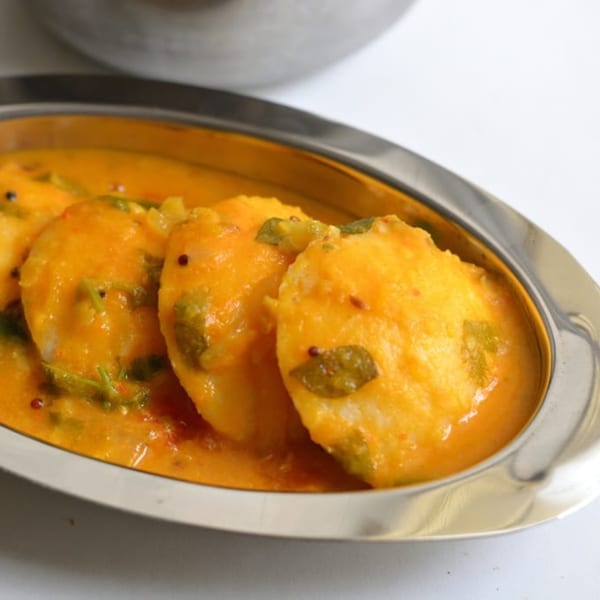 Three idlis covered in a rich orange sambar sauce, garnished with green herbs, in a silver dish.